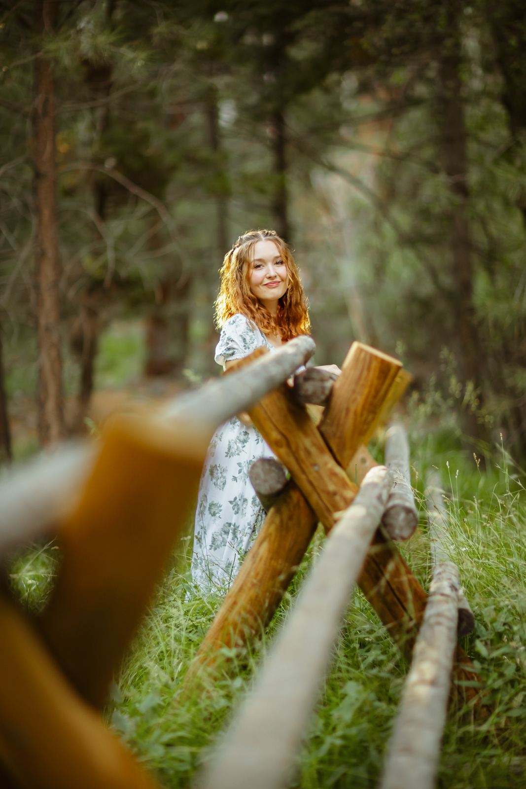 A young woman with wavy reddish hair stands in tall grass behind a rustic wooden log fence, smiling toward the camera. She wears a light floral print dress, framed by an out-of-focus evergreen forest in soft natural light.